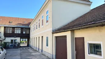 Ansicht eines Gebäudes mit hellem Putz, braunen Garagentoren und einem gepflasterten Hofbereich.