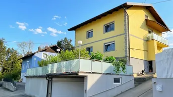 Gelbes Wohnhaus mit Satteldach, Balkon und integrierter Garage, umgeben von viel Grün unter blauem Himmel.