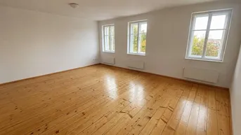 Heller, leerer Raum mit breiten Holzplankenböden, weißen Wänden und drei Fenstern mit Blick ins Grüne.