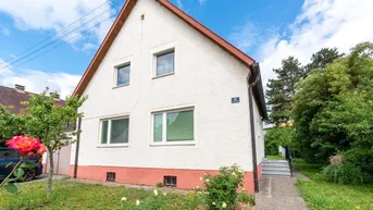 Vorderansicht eines Einfamilienhauses mit weißer Fassade und rotem Dach in einer grünen Umgebung.