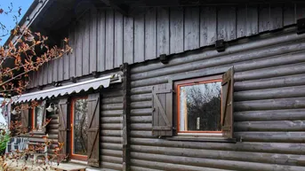 Dunkle Holzfassade eines Blockhauses mit Fenstern und einer Tür, umgeben von kahlen Ästen.