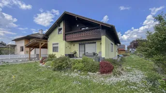 Hausfassade mit Balkon, Terrasse und gepflegtem Gartenbereich unter blauem Himmel.