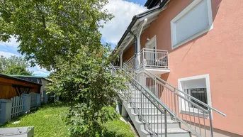 Seitenansicht des Hauses mit Außentreppe, die zu einem oberen Balkon führt, umgeben von Grün.