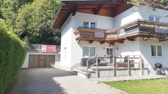 Hausfassade im alpinen Stil mit Holzdetails, einer Terrasse und einer gepflasterten Auffahrt zur Garage.