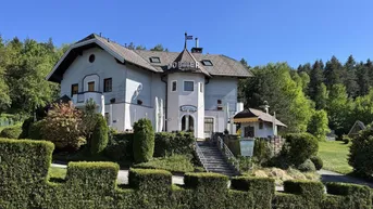 Großes Gebäude mit traditioneller Architektur, umgeben von gepflegten Hecken und grüner Landschaft unter blauem Himmel.