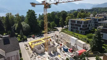 Baustelle mit Kran und im Bau befindlichem Gebäude, umgeben von Natur mit See- und Bergblick.