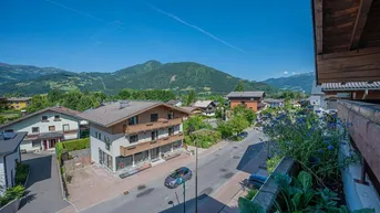 Blick von einem Balkon über eine ländliche Straße mit Häusern und grünen Bergen im Hintergrund.