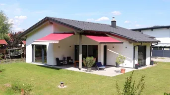 Modernes Einfamilienhaus mit großer Terrasse, roter Markise und gepflegtem Garten unter blauem Himmel.