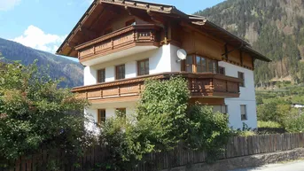 Traditionelles Haus im alpinen Stil mit Holzbalkonen und weißer Fassade, umgeben von Bergen.