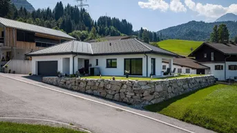 Modernes Einfamilienhaus mit weißer Fassade, dunklem Dach und integrierter Garage in alpiner Umgebung.