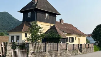 Historisches Gebäude mit markantem Turm und Satteldach, umgeben von einer Steinmauer und Holzzaun.