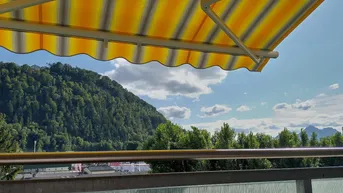 Blick vom Balkon mit gelber Markise auf bewaldete Berge und weite Landschaft.