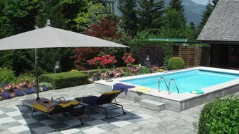 Gepflegter Poolbereich mit Sonnenliegen, Sonnenschirm und üppiger Gartenlandschaft.