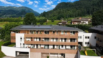 Modernes Mehrfamilienhaus mit Balkonen, Solaranlage und Spielplatz vor alpiner Bergkulisse.