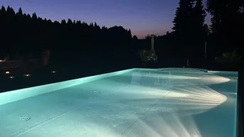 Beleuchteter Außenpool bei Dämmerung mit ruhiger Waldkulisse im Hintergrund und klarem Wasser.