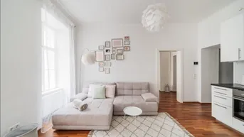 Heller Wohnbereich mit beigem Sofa, Holzboden und integrierter Küchenzeile, ideal für offenes Wohnen.