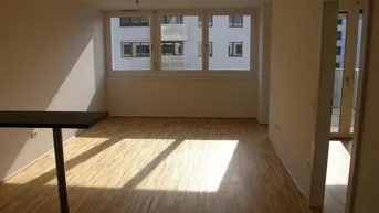 Helles, leeres Wohnzimmer mit Parkettboden und großen Fenstern, die auf ein gegenüberliegendes Gebäude blicken.