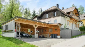 Traditionelles Haus mit großem Holzcarport und Stellplätzen, umgeben von grüner Landschaft.