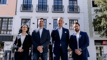 Moderne weiße Fassade eines Gebäudes mit schwarzen Balkonen und vier Personen davor.