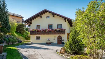 Traditionelles Haus mit Blumenbalkon und gepflasterter Auffahrt in ländlicher Umgebung.