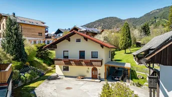 Hausfassade mit Balkon und Carport, umgeben von grüner Natur und Bergen.