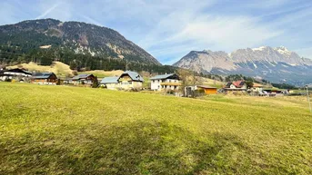 Weitläufige grüne Wiesen mit mehreren Häusern und beeindruckendem Bergpanorama im Hintergrund.
