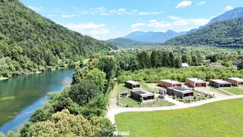 Luftaufnahme einer Siedlung moderner Häuser am Flussufer mit Bergblick und viel Grün.