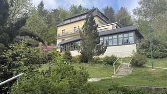 Mehrstöckiges Gebäude in Hanglage, umgeben von üppiger Vegetation und Bäumen unter blauem Himmel.