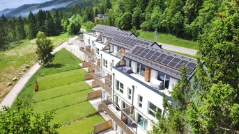 Moderne Reihenhäuser mit Photovoltaikanlagen auf den Dächern, eingebettet in eine grüne Berglandschaft.