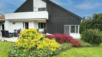 Moderne Hausfassade mit Terrasse, Garten und farbenfrohen Sträuchern unter blauem Himmel.