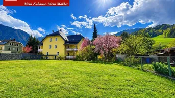 Gelbes Mehrfamilienhaus mit Balkonen und großem Gartenbereich vor einer Bergkulisse unter blauem Himmel.