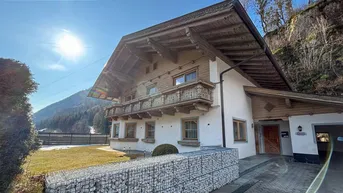 Traditionelles Haus im alpinen Stil mit Holzbalkonen und Gabionenmauer im Vorgarten, umgeben von Bergen.