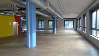 Großer, offener Gewerberaum mit hohen Decken, sichtbaren Lüftungsrohren und großen Fenstern, die viel Tageslicht hereinlassen.