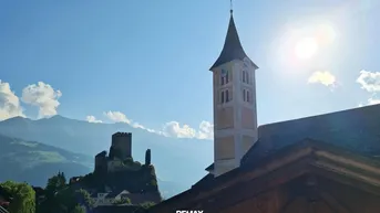 Panoramablick auf eine historische Kirche, eine Burgruine und die umliegende Berglandschaft unter strahlendem Himmel.
