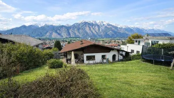 Haus mit Garten und Trampolin, umgeben von grüner Landschaft und schneebedeckten Bergen im Hintergrund.