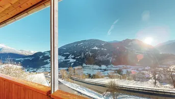 Sonniger Ausblick von einem Holz-Balkon auf eine verschneite Berglandschaft und eine Straße.