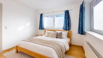 Helles Schlafzimmer mit Holzbett, Parkettboden und blauen Vorhängen, gut beleuchtet durch große Fenster.