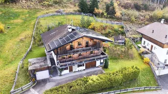 Luftaufnahme eines traditionellen Holzhauses mit Solaranlage, umgeben von Wald und Wiesen.
