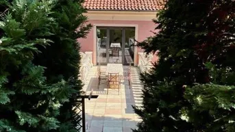 Blick auf eine sonnige Steinterrasse mit Gartenmöbeln vor der rosafarbenen Villa, umgeben von hohen Bäumen.