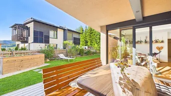 Gemütliche Terrasse mit Holztisch und Bank, umgeben von einem gepflegten Garten und modernen Häusern.
