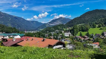 Panoramablick auf ein malerisches Dorf im Tal, umgeben von grünen Hügeln und majestätischen Bergen unter blauem Himmel.