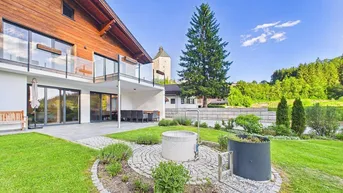 Hausfassade mit Holzverkleidung, großer Terrasse und gepflegtem Garten mit Blick auf die Landschaft.