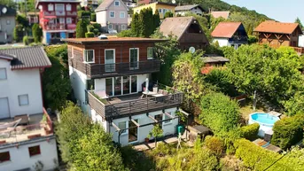 Modernes Haus mit Holzfassade, großer Terrasse, Balkon und Garten mit Pool auf einem grünen Hügel.