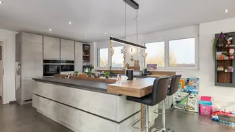 Moderne Küche mit großer Kücheninsel, integrierten Geräten und viel Tageslicht durch große Fenster.