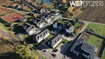 Luftaufnahme einer Wohnsiedlung mit Einfamilienhäusern, Grünflächen, einem Teich und Sportanlagen in ländlicher Umgebung.