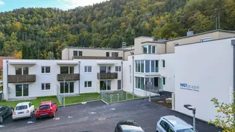 Modernes Mehrfamilienhaus mit weißen Fassaden, Balkonen und Parkplätzen vor bewaldetem Hügel.