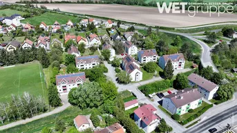 Luftaufnahme einer Wohnsiedlung mit Häusern, einigen davon mit Solaranlagen, umgeben von viel Grün und Feldern.