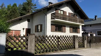 Vorderansicht des Hauses mit Balkon, Holzfensterläden und einem Lattenzaun unter blauem Himmel.