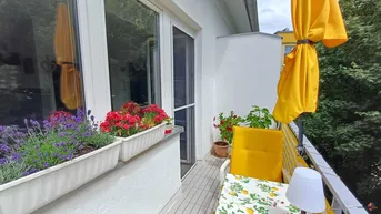 Heller Balkon mit gelbem Sonnenschirm, Blumenkästen und Holzboden, ideal zum Entspannen im Freien.