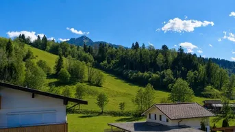 Panoramablick auf grüne Hügel und bewaldete Berge mit traditionellen Häusern unter blauem Himmel.